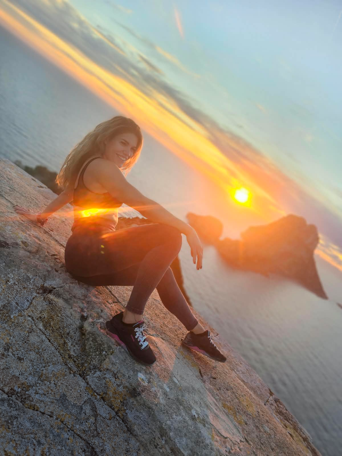 Simone sitzt lächelnd auf einem Felsen am Meer bei Sonnenuntergang